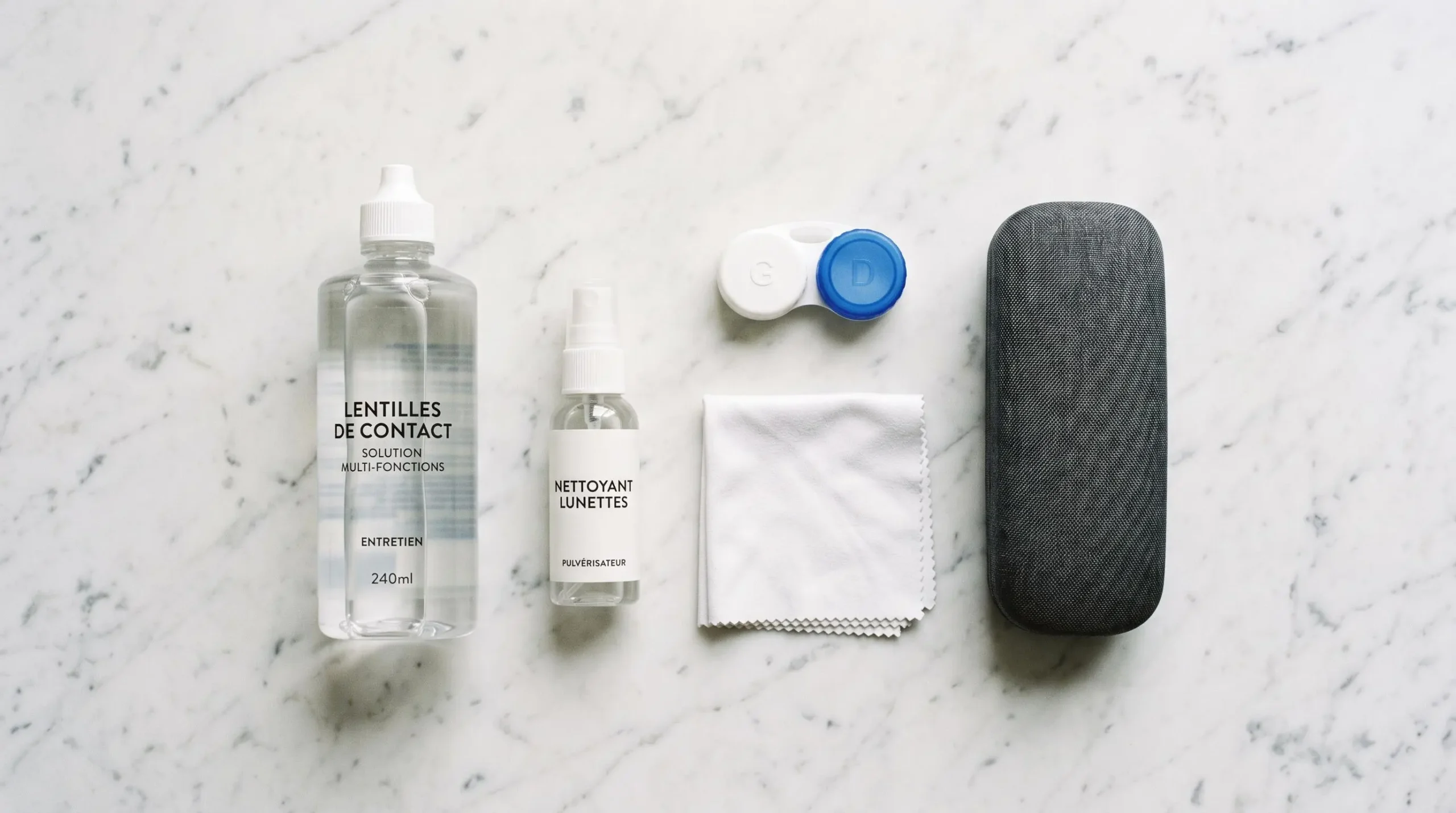 Vue du dessus d'un ensemble de produits d'entretien pour lunettes et lentilles disposés sur une surface blanche : solutions, étuis et chiffon microfibre