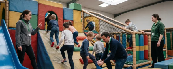 Trois adultes surveillent un groupe d'enfants jouant sur une structure gonflable et en mousse dans un parc indoor.